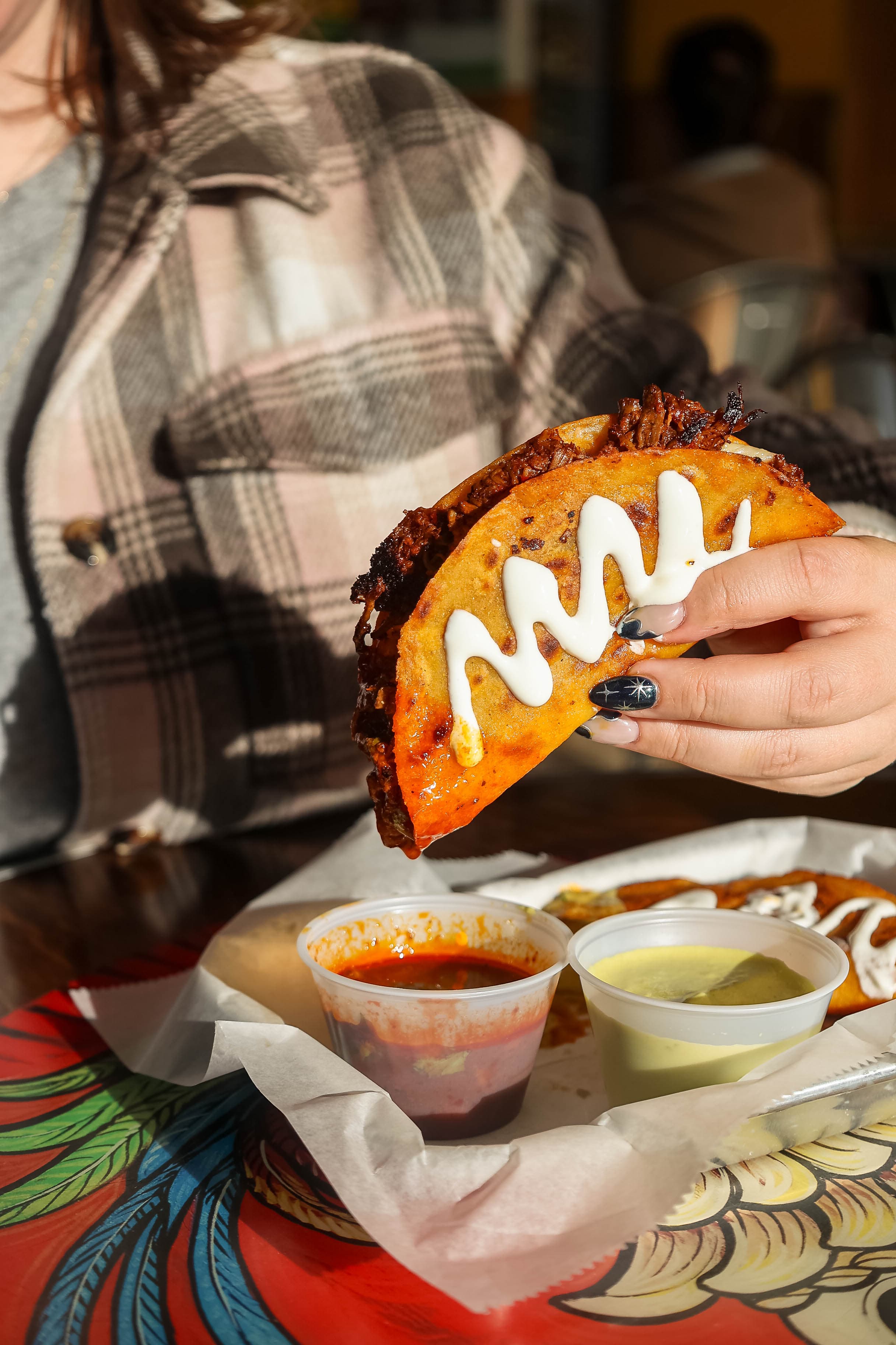 Close-up of a crispy birria taco with crema drizzle, held above a tray with red and green salsa.