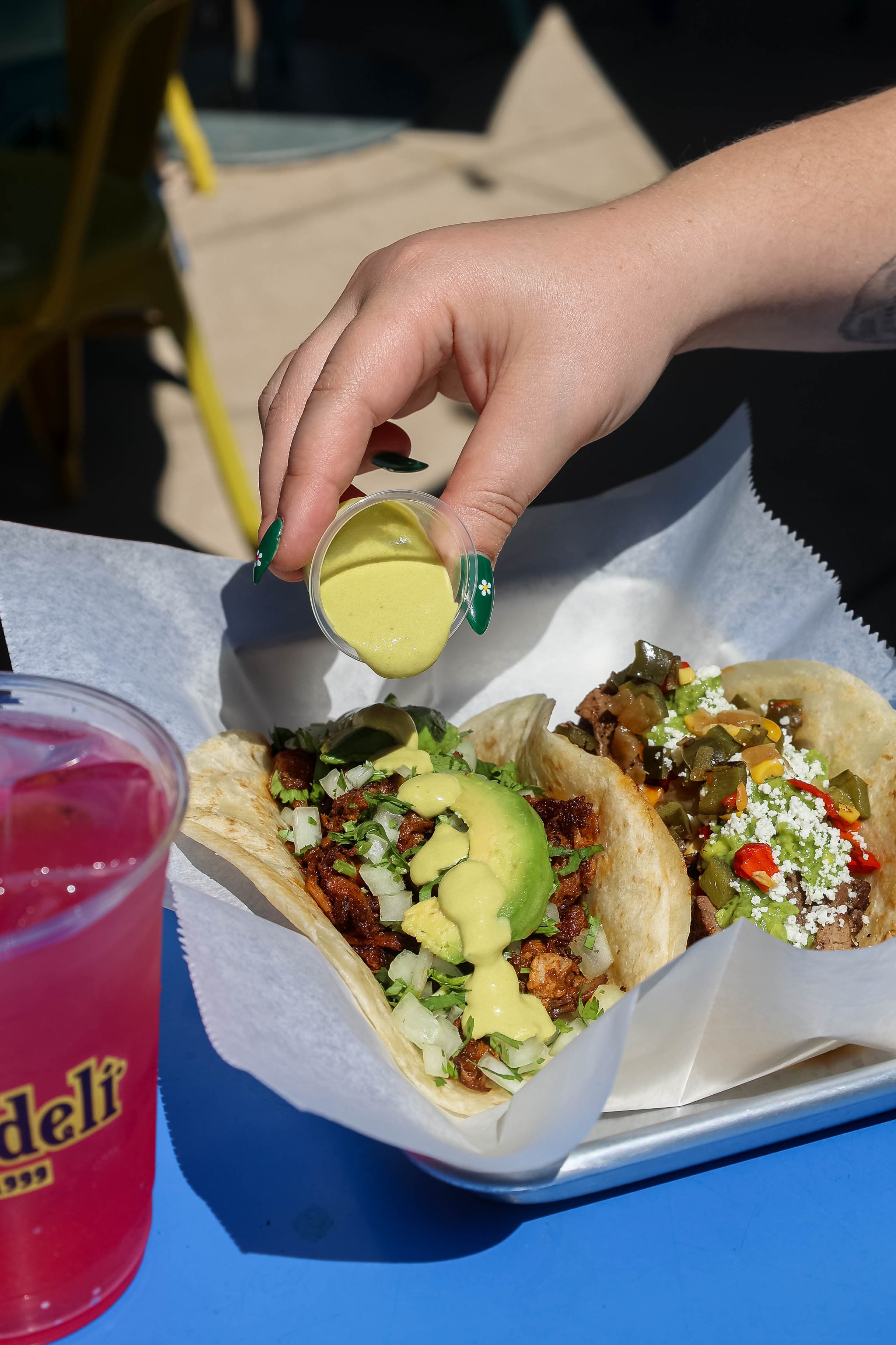 A person pours creamy green salsa from a small cup onto a taco topped with shredded meat, avocado slices, diced onions, and cilantro. Another taco with grilled beef, guacamole, queso fresco, and sautéed peppers is also visible on the tray. A bright pink drink is on the table beside them